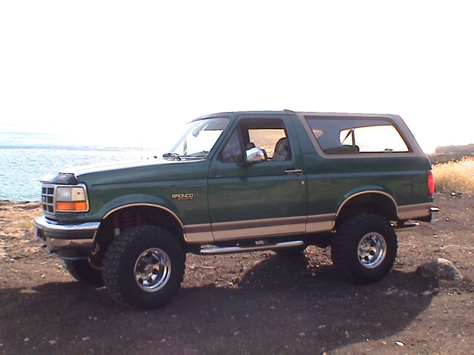 Green lifted Ford Bronco on chrome wheels and mud tires parked near a shoreline