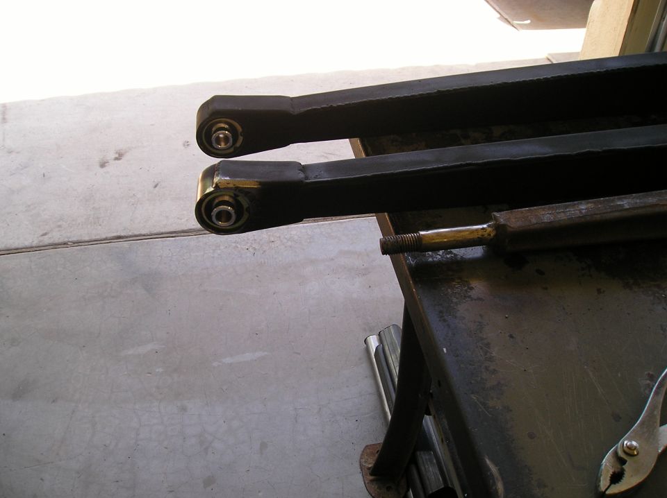 Two black suspension arms with bushings on a workbench in a garage