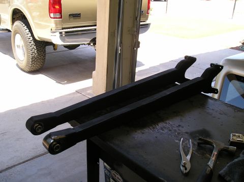 1993 Ford Bronco - Two black control arms are laid out on a workbench, with a portion of a tan Ford truck visible behind them.