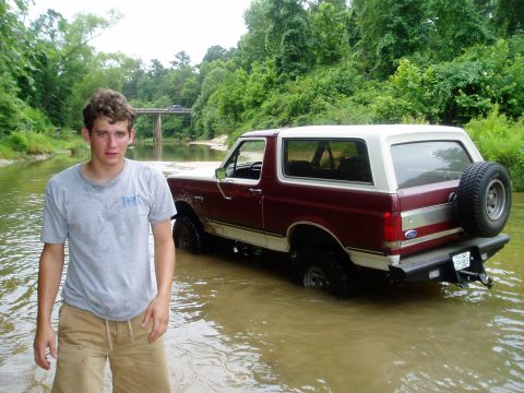 1990 Ford Bronco - Man and 1990 Ford Bronco partially submerged in a creek, with a bridge in the background.