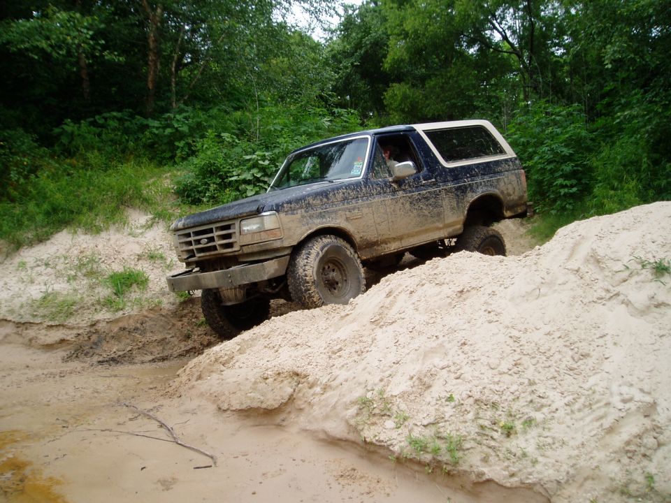 Mud-covered dark blue and white Ford Bronco driving off-road through sand and mud