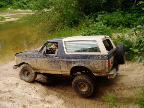 1990 Ford Bronco - Muddy 1990 Ford Bronco navigating a dirt trail next to a body of water.