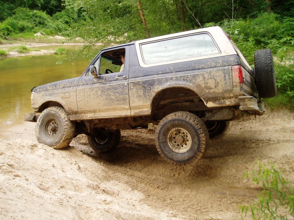 Muddy SUV driving on a dirt path near water. It has a spare tire mounted on the back