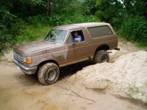 1990 Ford Bronco - A muddy brown Ford Bronco navigates a sandy off-road trail with a driver behind the wheel.