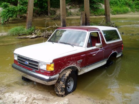 1990 Ford Bronco - This Ford Bronco is navigating a muddy, shallow creek bed, with concrete bridge supports visible in the background.