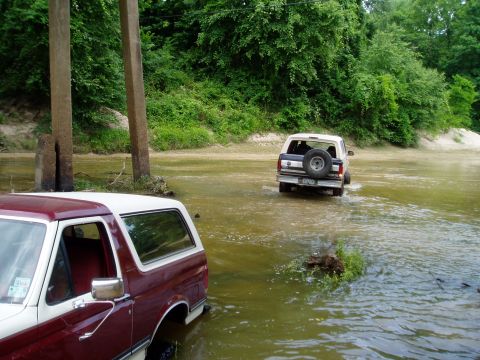 1990 Ford Bronco - Two Ford Broncos navigate a shallow river crossing surrounded by lush green trees.