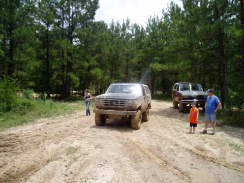 1990 Ford Bronco - Two Ford Broncos are parked on a muddy track in a wooded area with several people nearby.