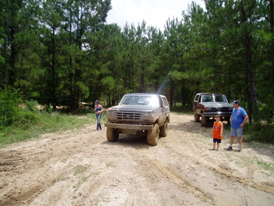 Two muddy Ford Broncos parked on a dirt track with people standing around. Trees in background