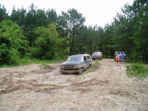 1990 Ford Bronco - Several SUVs are seen off-roading on a dirt track surrounded by trees, with one Bronco stuck in the mud.