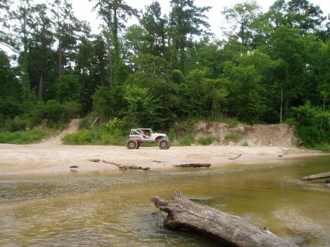 1990 Ford Bronco - An off-road buggy is parked on a sandy bank next to a wooded area and a river.
