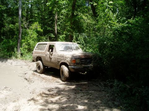 1990 Ford Bronco - This SUV is covered in mud and appears to be stuck in a muddy trail surrounded by dense foliage.