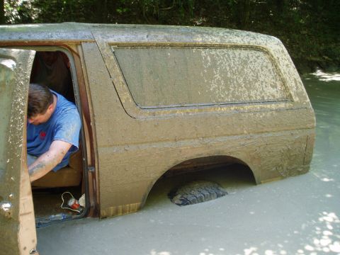 1990 Ford Bronco - A mud-covered SUV is submerged up to its windows in a muddy water hole, with a man in a blue t-shirt leaning out of the open front door rea…