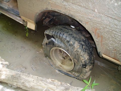 1990 Ford Bronco - A mud-caked truck sits axle-deep in a brown water hole with a badly damaged rear tire, the sidewall is torn open and the mud-terrain tread…