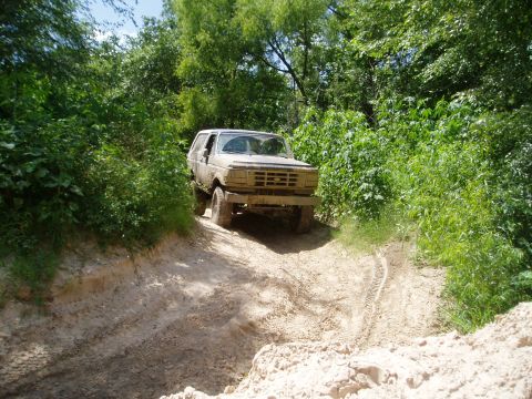 1990 Ford Bronco - A dusty Ford Bronco navigates a muddy trail surrounded by lush green foliage.