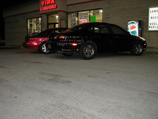 Black sedan parked next to a red car in front of a liquor store at night. Supermotors.net logo visible