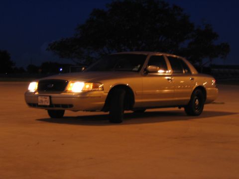 2000 Ford Crown Victoria - A gold 2000 Ford Crown Victoria sedan is parked on a concrete surface at night with its headlights on.