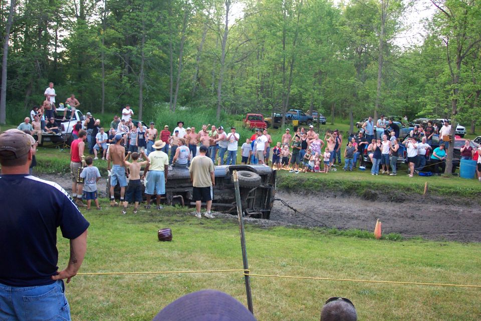 Crowd gathered around a pickup rolled on its side in a muddy bog pit