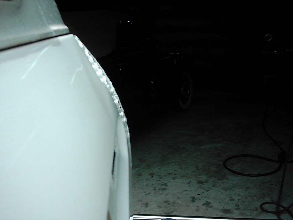White vehicle fender with visible damage along the edge, showing a dark interior and another vehicle in the background