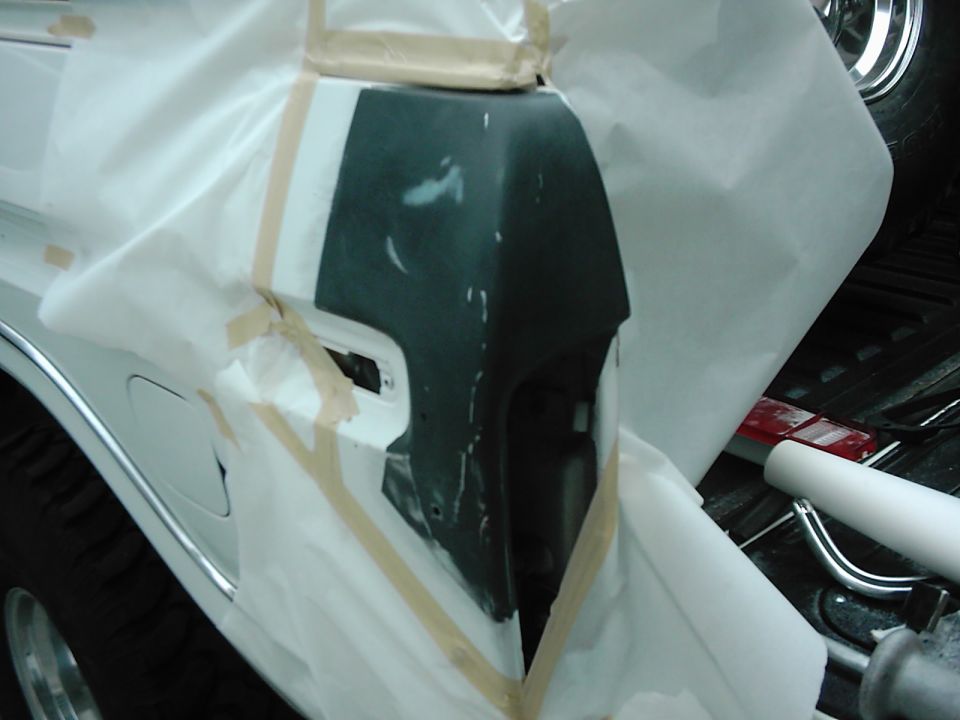Vehicle rear bumper and fender area covered in masking paper and tape, with primer applied to the bumper. A Supermotors.net watermark is visible