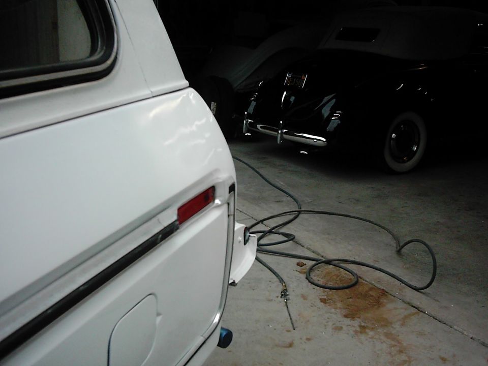 White SUV rear corner with taillight and fuel door visible. Black classic car in background. Hoses on floor. Supermotors.net logo