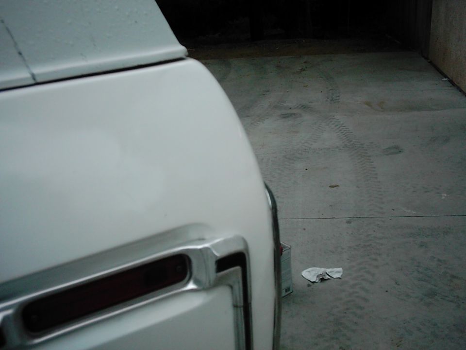 Close-up of the rear corner of a white vehicle, showing part of a taillight and chrome trim. The ground has tire tracks