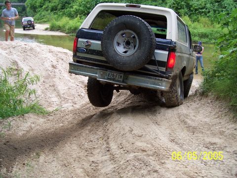 1987 Ford Bronco - A Ford Bronco navigates a sandy trail near a body of water, with another vehicle visible in the background.