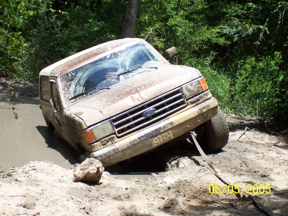 Mud-covered brown Ford Bronco stuck nose-down in a mud pit with a recovery strap attached, dated 06/05/2005