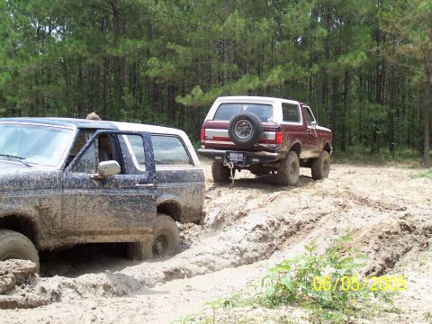 1987 Ford Bronco - Two Ford Broncos navigate a muddy off-road trail, with one vehicle deeply stuck in the mire.