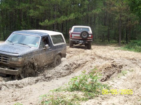 1987 Ford Bronco - Two Ford Broncos navigate a muddy off-road trail, with one vehicle stuck deep in the mire.