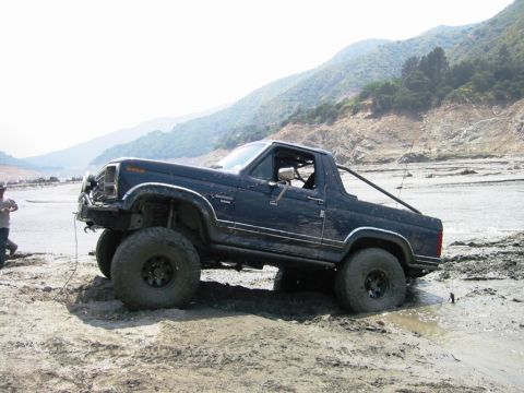 1985 Ford Bronco - A modified Ford Bronco is stuck in a muddy, off-road environment with a person attempting to pull it out.