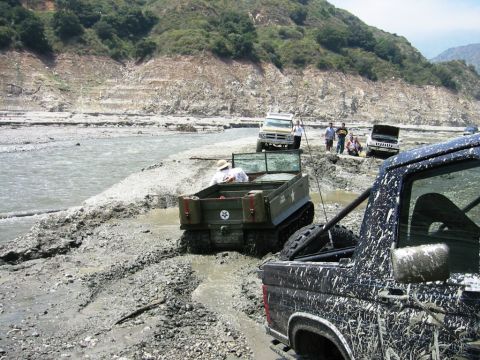 1985 Ford Bronco - Several vehicles navigate a muddy, rocky terrain near a river, with a black pickup truck in the foreground.