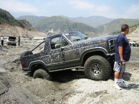 1985 Ford Bronco - This SUV is deep in the mud, with a man observing the situation. Other vehicles are present in the background.