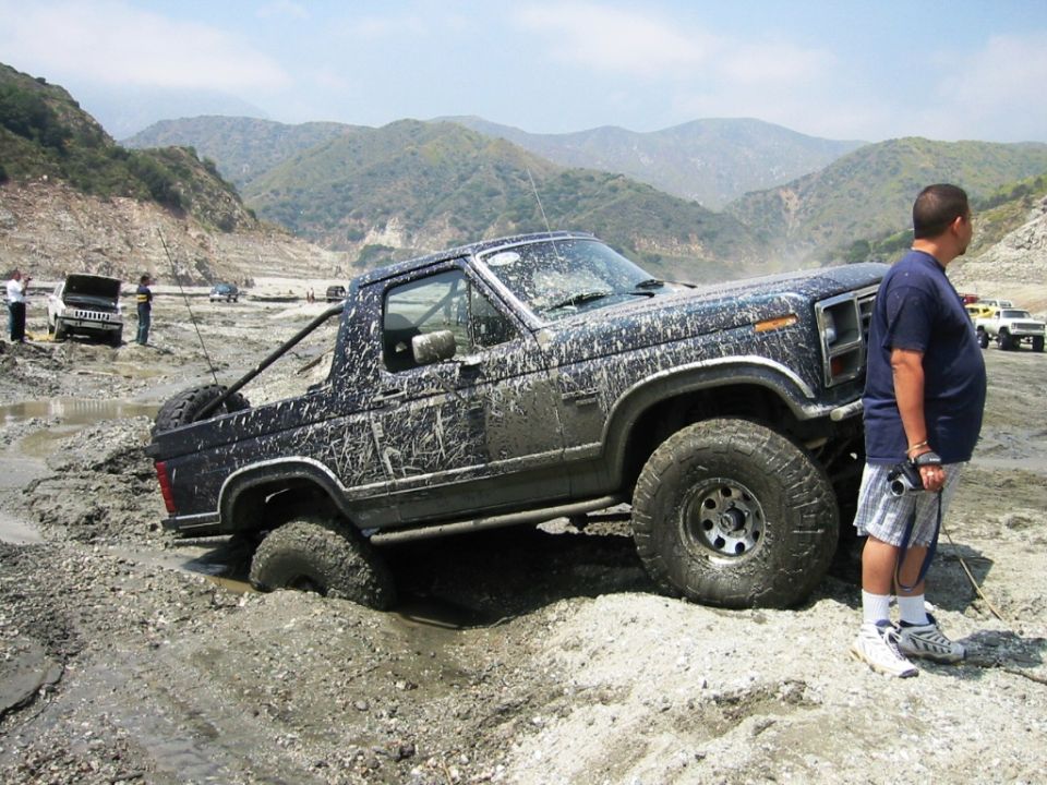 Mud-covered dark blue Ford Bronco stuck in mud, man standing beside it in mountains