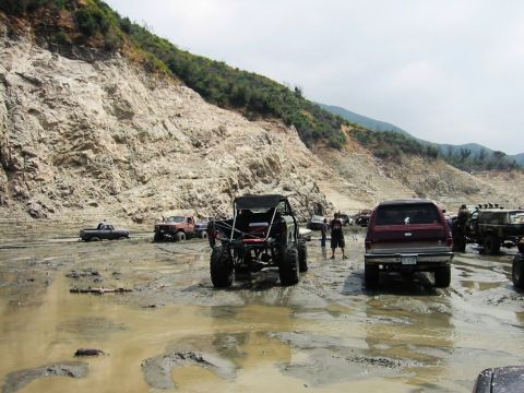 1985 Ford Bronco - Several off-road vehicles are parked in a muddy area with a large, textured cliff in the background.