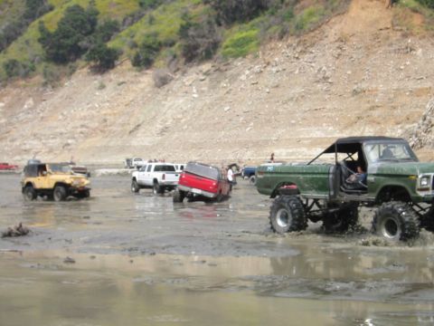 1985 Ford Bronco - Several off-road vehicles navigate a muddy terrain, with a prominent green lifted pickup truck in the foreground.