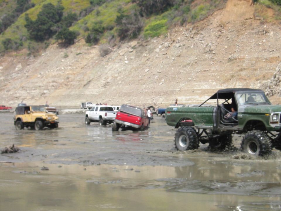 Green lifted Ford Bronco on big mud tires plowing through a muddy off-road basin with other 4x4s.