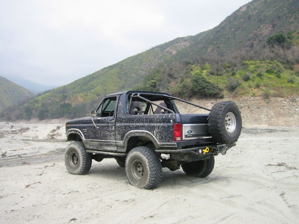 Rear 3/4 view of lifted topless dark Ford Bronco with roll cage and mud tires on sand