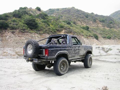 1985 Ford Bronco - This modified Ford Bronco is shown from the rear, equipped with an external spare tire and roll cage, on a sandy, outdoor location.
