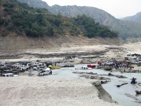 1985 Ford Bronco - A large gathering of vehicles, including trucks and SUVs, are situated in a dry riverbed with a wooded hillside.