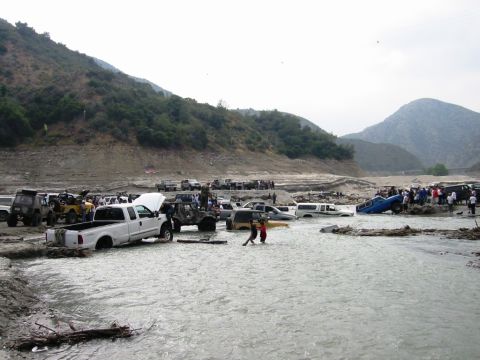 1985 Ford Bronco - Multiple off-road vehicles are submerged in a shallow river, with onlookers gathered on the banks.