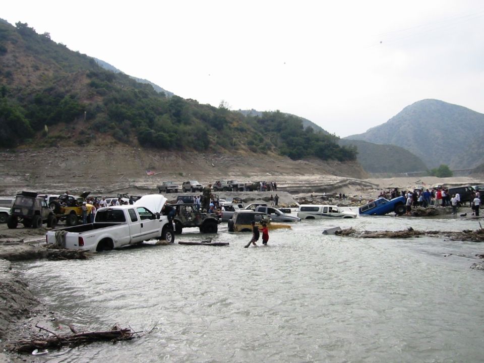 Off-road trucks and Jeeps crossing a muddy river in a mountain canyon with people wading