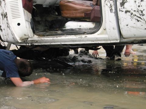 1985 Ford Bronco - People are reaching under a muddy truck chassis in shallow water, with the interior visible.