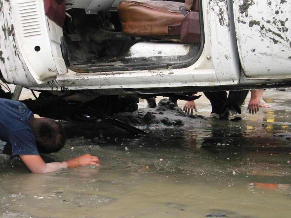 Muddy white vehicle stuck in floodwater with people working around it