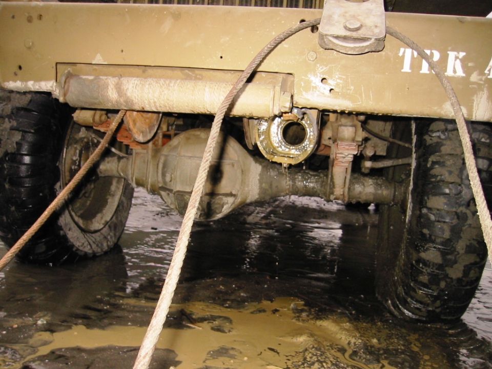 Rear axle and differential of a tan military-style vehicle over muddy floor with winch cable and pintle hitch