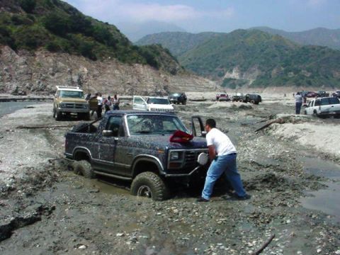 1985 Ford Bronco - A 1985 Ford Bronco is stuck in the mud while a man attempts to push it free, with other vehicles visible in the background.