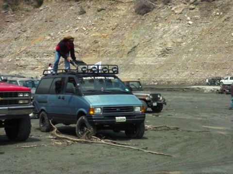1985 Ford Bronco - A blue van with a roof rack is parked on a dirt lot with several other vehicles and a person standing on its roof.