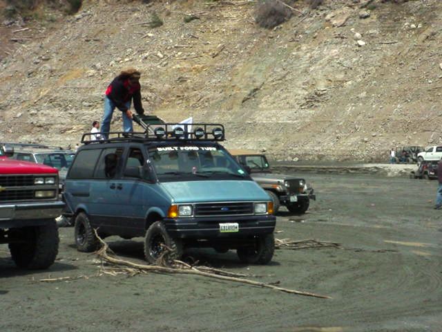 Lifted blue Ford Aerostar van with roof rack in dirt lot, person on roof