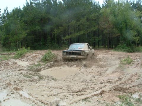 1993 Ford Bronco - A tan pickup truck navigates a muddy off-road course, kicking up spray as it goes. The grille suggests a 1973-1980 Chevrolet or GMC C/K ser…