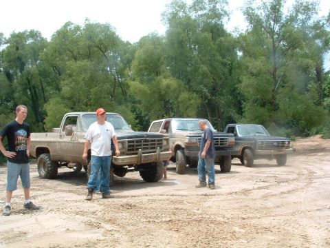 1993 Ford Bronco - Several off-road vehicles are gathered on a sandy area with trees in the distance.