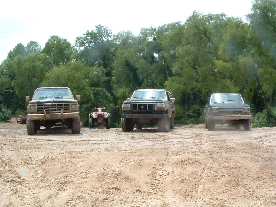 Three trucks and two ATVs parked on a dirt track with trees in the background